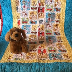 K&ouml;nnte beinhalten: Eine gelbe, blaue und rote Decke mit einem Muster aus Hunden und den Worten "Dogs & Suds" und "Woof". Ein braunes Pl&uuml;schhundspielzeug sitzt auf der Decke.