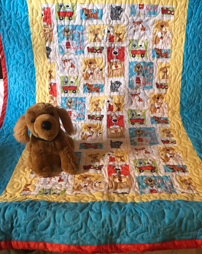 K&ouml;nnte beinhalten: Eine gelbe, blaue und rote Decke mit einem Muster aus Hunden und den Worten "Dogs & Suds" und "Woof". Ein braunes Pl&uuml;schhundspielzeug sitzt auf der Decke.