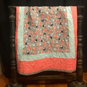 Op de afbeelding: Een koraalroze en lichtblauwe babydeken met een grijze en oranje vossenprint. De deken is gestikt met een swirl-patroon en heeft een grijze rand.