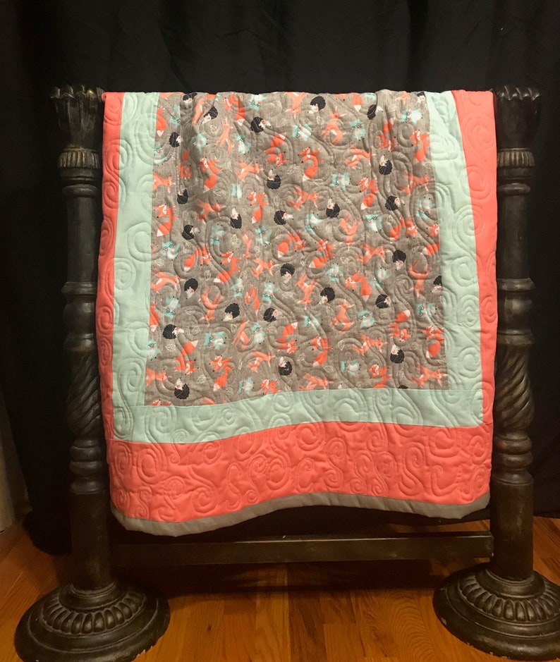 Op de afbeelding: Een koraalroze en lichtblauwe babydeken met een grijze en oranje vossenprint. De deken is gestikt met een swirl-patroon en heeft een grijze rand.