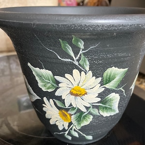 May include: A dark gray flower pot with a textured surface. The pot is decorated with hand-painted white daisies with yellow centers and green leaves. The floral design is set against a dark background.