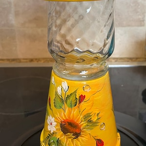 May include: A yellow painted ceramic jar with a glass insert and a yellow lid. The jar is decorated with a sunflower, white flowers, and green leaves.
