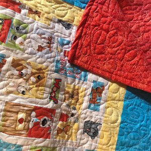 K&ouml;nnte beinhalten: Eine bunte Quiltdecke mit Patchwork-Muster, die verschiedene Bilder von Hunden zeigt. Die Decke hat einen blauen, gelben und wei&szlig;en Hintergrund mit roten Akzenten. Die Decke ist aus Baumwollstoff gefertigt und mit einem einfachen Steppmuster gesteppt. Die Decke ist in der H&auml;lfte gefaltet und die R&uuml;ckseite ist einfarbig rot.
