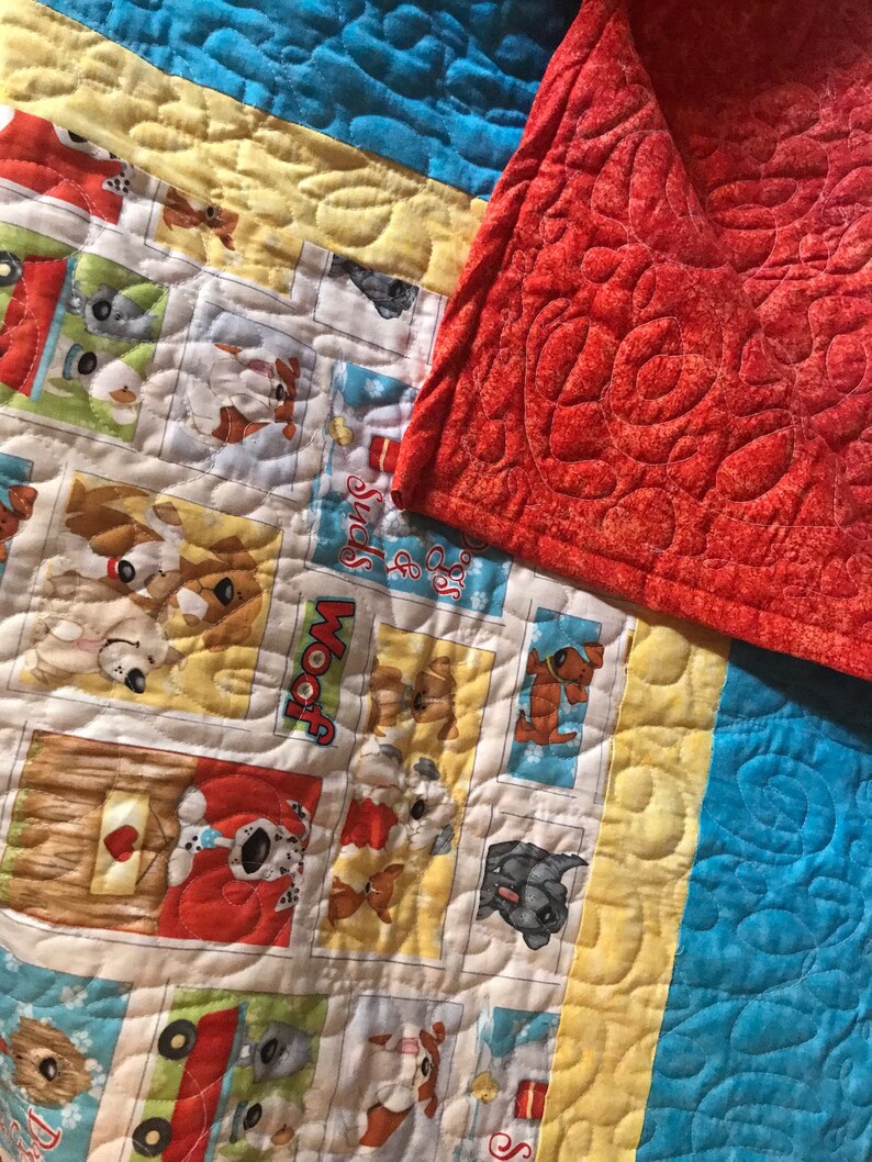 K&ouml;nnte beinhalten: Eine bunte Quiltdecke mit Patchwork-Muster, die verschiedene Bilder von Hunden zeigt. Die Decke hat einen blauen, gelben und wei&szlig;en Hintergrund mit roten Akzenten. Die Decke ist aus Baumwollstoff gefertigt und mit einem einfachen Steppmuster gesteppt. Die Decke ist in der H&auml;lfte gefaltet und die R&uuml;ckseite ist einfarbig rot.