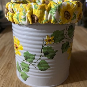 May include: A white metal can with a sunflower design painted on it. The can is decorated with a yellow and white fabric band with a sunflower pattern.
