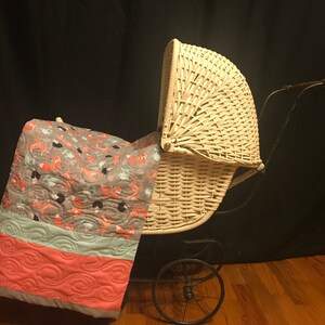 Op de afbeelding: Een witte rieten kinderwagen met een zwart metalen frame en wielen. Een grijs, roze en blauw gepatroneerd dekbed is over de kinderwagen gelegd.