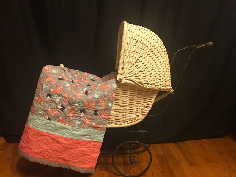 Op de afbeelding: Een witte rieten kinderwagen met een zwart metalen frame en wielen. Een grijs, roze en blauw gepatroneerd dekbed is over de kinderwagen gelegd.