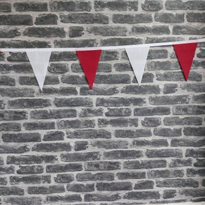 May include: A decorative bunting featuring alternating red and white triangular flags. The flags are attached to a white ribbon. The background is a grey brick wall, providing a contrasting backdrop.