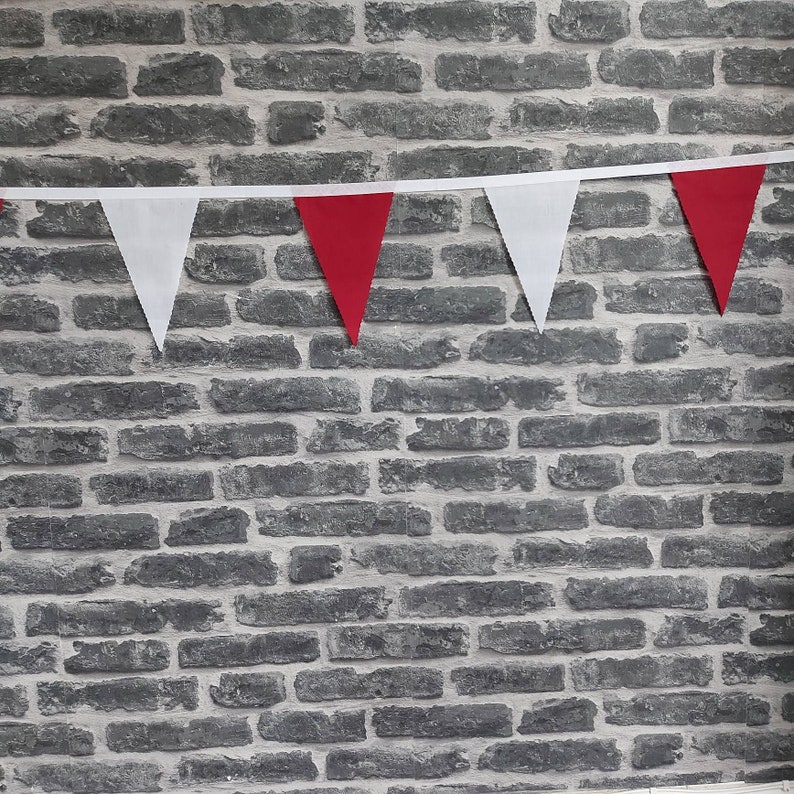 May include: A decorative bunting featuring alternating red and white triangular flags. The flags are attached to a white ribbon. The background is a grey brick wall, providing a contrasting backdrop.