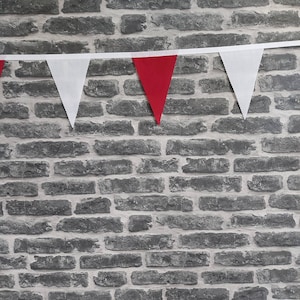 May include: A decorative bunting with alternating white and red triangular flags, suspended against a grey brick wall. The flags are made of fabric and are attached to a white ribbon. The wall has a textured finish.