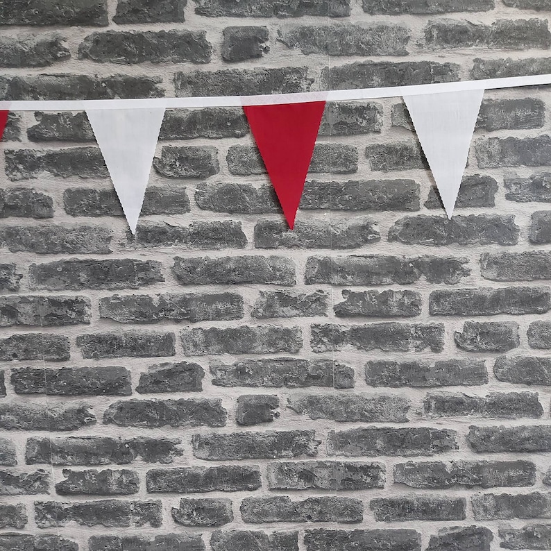 May include: A decorative bunting with alternating white and red triangular flags, suspended against a grey brick wall. The flags are made of fabric and are attached to a white ribbon. The wall has a textured finish.