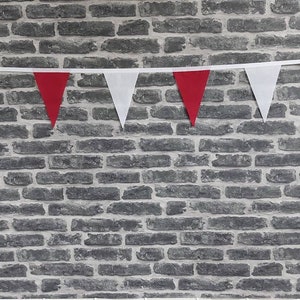 May include: A decorative bunting of alternating red and white triangular flags against a grey brick wall. The flags are strung on a white ribbon, creating a festive display. The brick wall provides a textured backdrop.