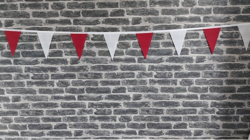 May include: A decorative bunting of alternating red and white triangular flags against a grey brick wall. The flags are strung on a white ribbon, creating a festive display. The brick wall provides a textured backdrop.