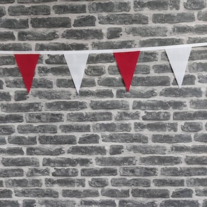 May include: A bunting of red and white triangular flags against a grey brick wall. The alternating red and white flags are attached to a white ribbon. The pennants are evenly spaced, creating a festive, decorative display.