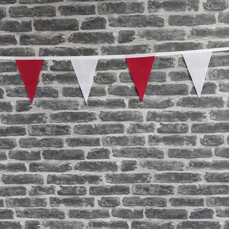 May include: A bunting of red and white triangular flags against a grey brick wall. The alternating red and white flags are attached to a white ribbon. The pennants are evenly spaced, creating a festive, decorative display.