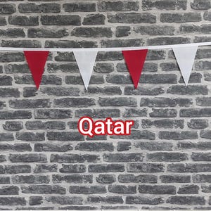 May include: A banner of red and white triangular flags against a grey brick wall. The word "Qatar" is displayed in red text with a white outline. The flags are attached to a white string.
