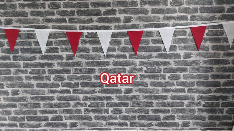 May include: A banner of red and white triangular flags against a grey brick wall. The word "Qatar" is displayed in red text with a white outline. The flags are attached to a white string.