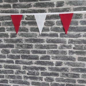 May include: A length of red and white triangular bunting suspended against a grey brick wall. The bunting is strung on a white ribbon. The red triangles alternate with white triangles.