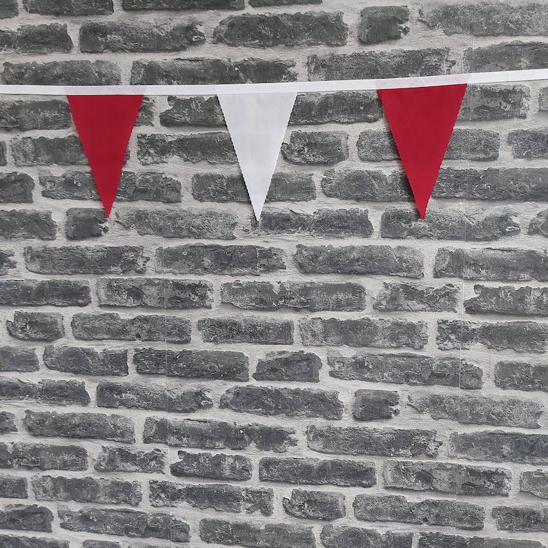 May include: A length of red and white triangular bunting suspended against a grey brick wall. The bunting is strung on a white ribbon. The red triangles alternate with white triangles.