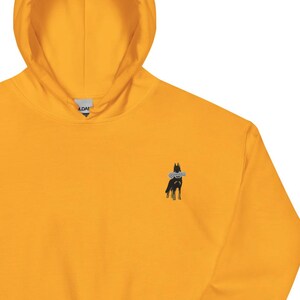 May include: Mustard yellow hoodie with a kangaroo pocket and a hood. The hoodie features a small embroidered design of a dog on the upper left chest. The hoodie is made of a soft material.