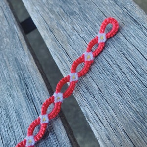 May include: A handmade bracelet featuring a chain-like design with red and white woven threads. Small white and yellow flower accents are evenly spaced along the bracelet. The bracelet is displayed on a weathered wooden surface.