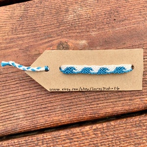 May include: A blue and white friendship bracelet with a wave pattern. The bracelet is displayed on a brown paper tag with the website address www.etsy.com/shop/SarasStudio26.