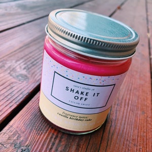 May include: A pink candle in a glass jar with a silver lid. The label says "sara's studio 26 Shake It Off Taylor Swift" and "fragrance notes: vanilla birthday cake".