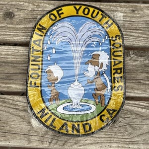May include: Oval patch with the words "FOUNTAIN OF YOUTH SQUARES NILAND CA" in a yellow border. The patch features a cartoon fountain with two figures and a blue and white background.