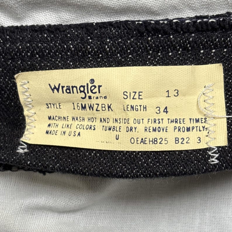 May include: A black fabric garment tag featuring the Wrangler brand logo, size 13, and length 34. The label includes style number 16MWZBK, care instructions for machine washing, and the text Made in USA, printed in black on a light beige background.