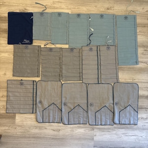 Può includere: Un assortimento di articoli in tessuto grigio e blu, tra cui sacchetti e tappetini piegati, disposti su una superficie di legno. Alcuni articoli presentano un logo circolare e dettagli cuciti. È presente anche un sacchetto blu navy con testo.