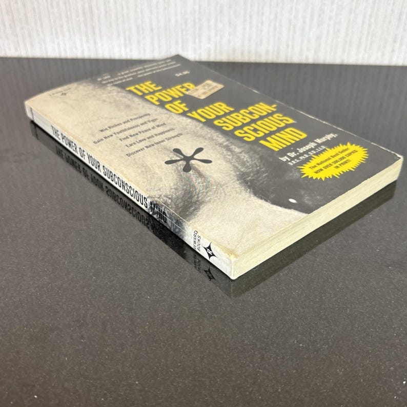 May include: A paperback book titled "The Power of Your Subconscious Mind" by Dr. Joseph Murphy. The cover features a black and white image of a face and the title in yellow and white. The book is on a dark surface.