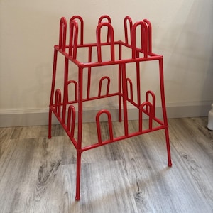 May include: A bright red metal boot rack featuring a square frame with four legs and multiple vertical U-shaped prongs extending upward from the top and sides, designed to hold footwear in an inverted position for storage and organization.