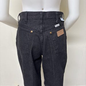 May include: Black denim jeans featuring a high-rise waist, belt loops, and a back yoke. The jeans include two back pockets with gold-tone metal rivets, a tan leather-like patch on the right pocket, and a small white Wrangler brand tag.