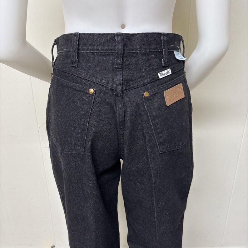 May include: Black denim jeans featuring a high-rise waist, belt loops, and a back yoke. The jeans include two back pockets with gold-tone metal rivets, a tan leather-like patch on the right pocket, and a small white Wrangler brand tag.
