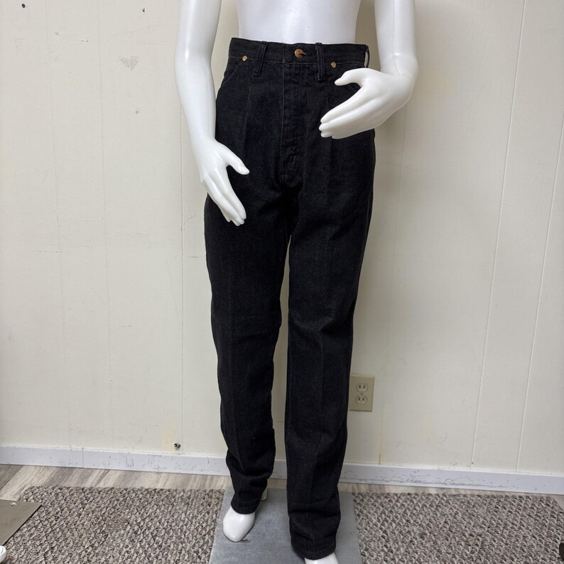 May include: High-waisted black denim jeans featuring a button fly closure, front pleats, and copper-toned rivets at the pockets. These straight-leg trousers are constructed from a dark, solid-coloured fabric with visible vertical stitching details.