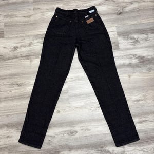 May include: A pair of high-waisted black denim jeans featuring a straight-leg cut, a button and zip fly closure, belt loops, and two back pockets. One back pocket displays a small rectangular brown leather patch with the Wrangler brand logo.