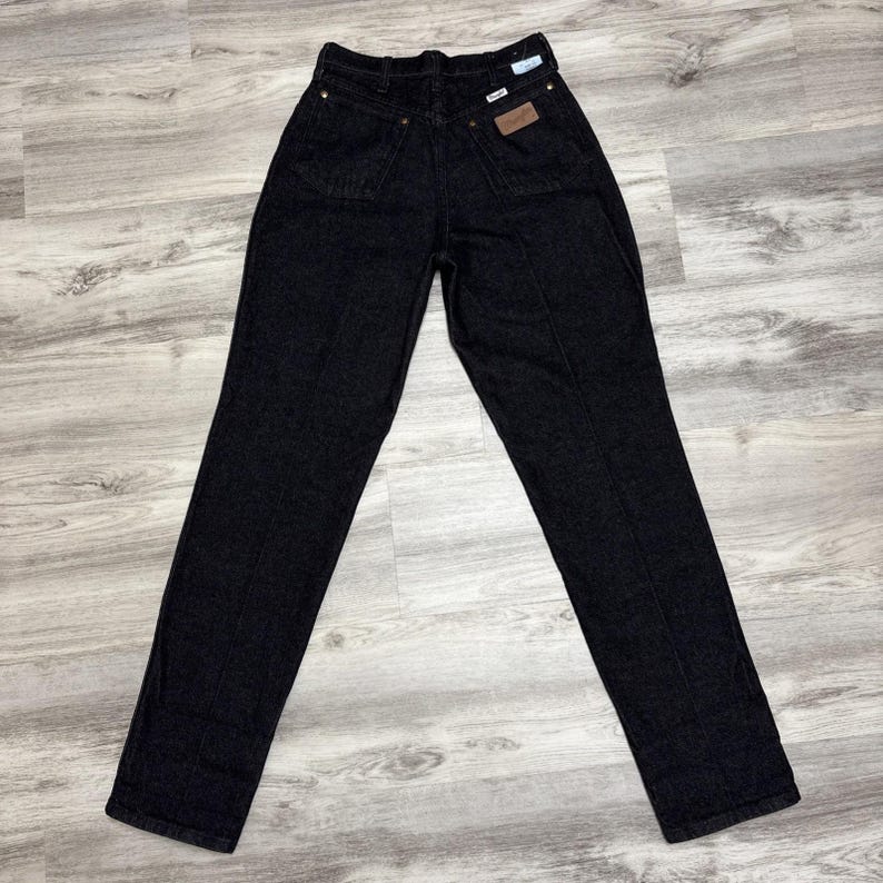 May include: A pair of high-waisted black denim jeans featuring a straight-leg cut, a button and zip fly closure, belt loops, and two back pockets. One back pocket displays a small rectangular brown leather patch with the Wrangler brand logo.