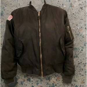 May include: A dark brown bomber jacket with a gold zip and a small American flag patch on the left shoulder. The jacket has a ribbed collar, cuffs, and waistband. It features two front pockets and a zipped pocket on the left sleeve.