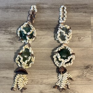 May include: Two decorative hanging ornaments. Each ornament features a clear green glass ball encased in a ring of small, white seashells. The ornaments are adorned with additional seashells and a cluster of brown shells.