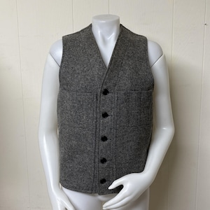 May include: A gray wool vest with a V-neck and a button-up front. The vest has two front pockets and is displayed on a mannequin. The vest is a neutral color and could be worn as a layering piece.