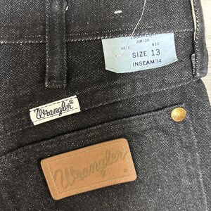 May include: Black denim jeans featuring a tan leather patch embossed with the Wrangler logo, a white fabric tag with the Wrangler brand name, and a light blue paper tag displaying the text JUNIOR, SIZE 13, and INSEAM 34 with a gold rivet.