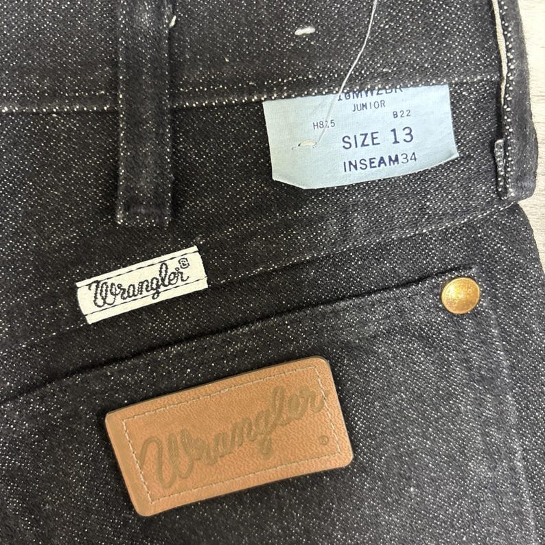 May include: Black denim jeans featuring a tan leather patch embossed with the Wrangler logo, a white fabric tag with the Wrangler brand name, and a light blue paper tag displaying the text JUNIOR, SIZE 13, and INSEAM 34 with a gold rivet.