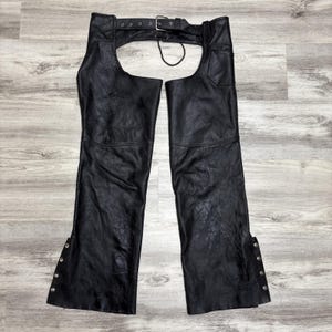May include: Black leather chaps with a belt and buckle closure at the waist. The chaps have a classic design with a textured finish and snap closures along the outer seams. The chaps are designed to be worn over pants.