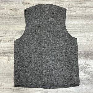 May include: A gray wool vest with a V-neck and a button closure. The vest is sleeveless and has a simple, classic design. The fabric has a textured appearance.