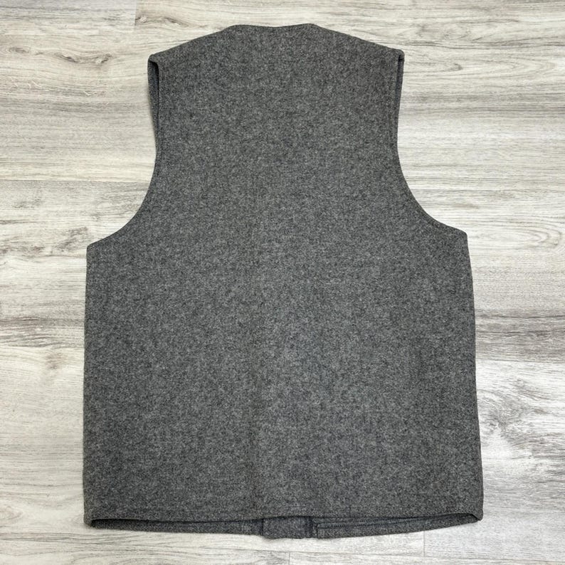 May include: A gray wool vest with a V-neck and a button closure. The vest is sleeveless and has a simple, classic design. The fabric has a textured appearance.