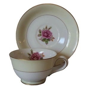 May include: A vintage teacup and saucer set. The cup and saucer are cream-colored with a gold trim. A pink rose is featured in the center of the saucer and inside the cup. The cup has a handle and sits on a small foot.