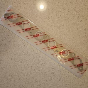 Könnte beinhalten: Eine weiße Verpackung mit einer silbernen Abgaskrümmerdichtung mit sechs Löchern. Die Dichtung trägt sechsmal den Schriftzug "TOYOTA".