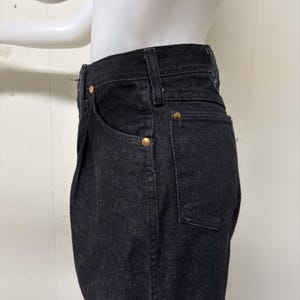May include: High-waisted black denim jeans featuring a classic five-pocket design with visible belt loops and metallic copper-toned rivets at the pocket corners. The fabric has a textured weave and a deep black colour throughout the garment.
