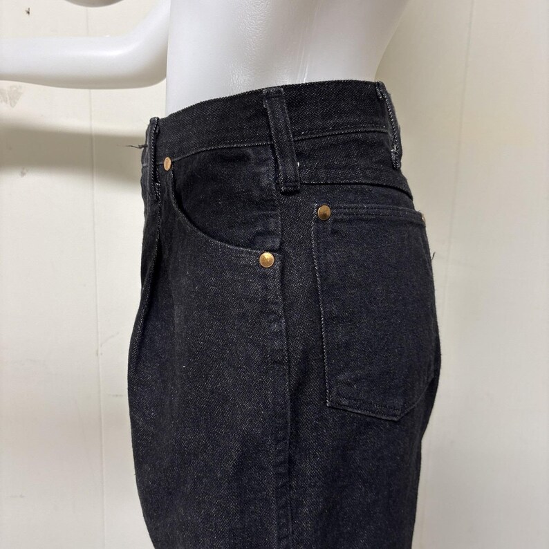 May include: High-waisted black denim jeans featuring a classic five-pocket design with visible belt loops and metallic copper-toned rivets at the pocket corners. The fabric has a textured weave and a deep black colour throughout the garment.