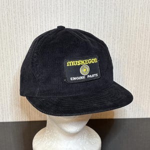 May include: Black corduroy baseball cap with a curved peak. A rectangular patch on the front displays the text "MUSKEGON ENGINE PARTS" in yellow and white, with a yellow circle design. The cap is displayed on a mannequin head.